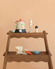 The Stir and Serve Soup Maker is displayed on a wooden shelf, featuring a toy stockpot, hand blender, pepper grinder, spoon, bag of beans, veggies, bowl, and spoon—all set against a beige background for imaginative play.