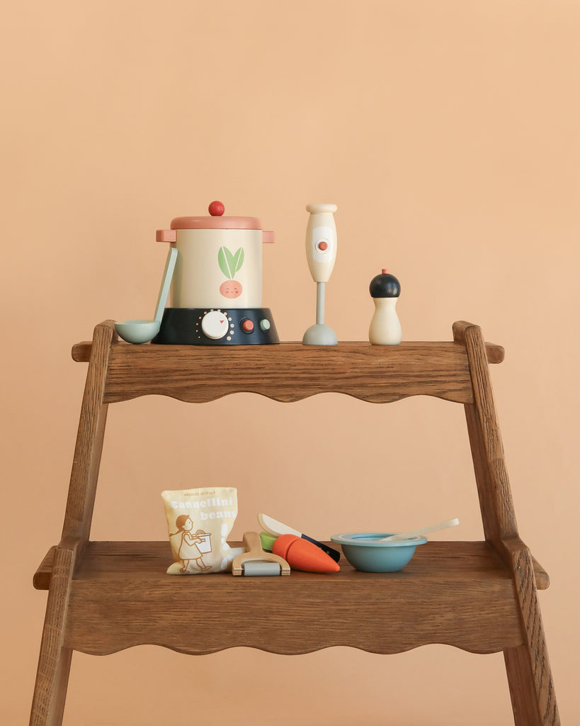 The Stir and Serve Soup Maker is displayed on a wooden shelf, featuring a toy stockpot, hand blender, pepper grinder, spoon, bag of beans, veggies, bowl, and spoon—all set against a beige background for imaginative play.