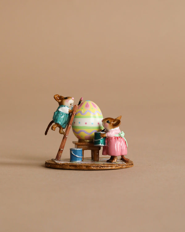 Two Wee Forest Folk Tiny Collectible mice in vibrant outfits paint a giant Easter egg—one on a ladder, one with a paint can—set on a table against a beige background. "Colossal Easter Coloring" is perfect for miniature fans.