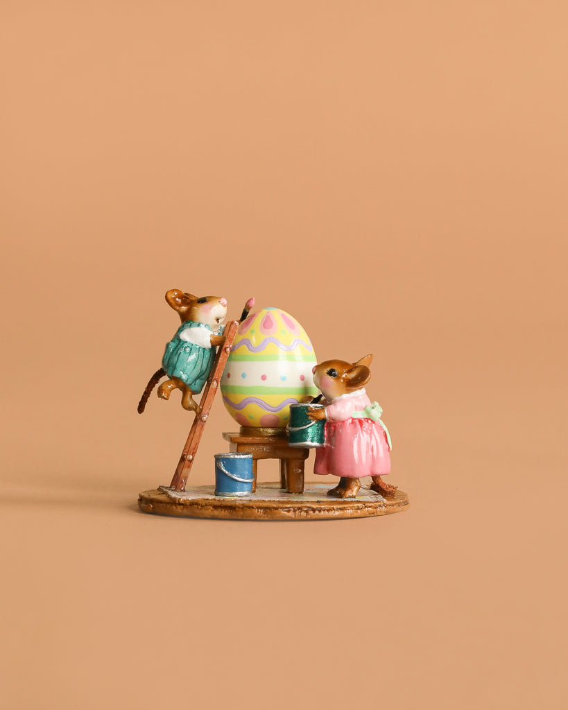 Two Wee Forest Folk mice in pastel outfits paint a big Easter egg—one on a ladder, one by paint cans—against a light brown background. The Wee Forest Folk Tiny Collectible - Colossal Easter Coloring is ideal for collectible mouse figurine fans.