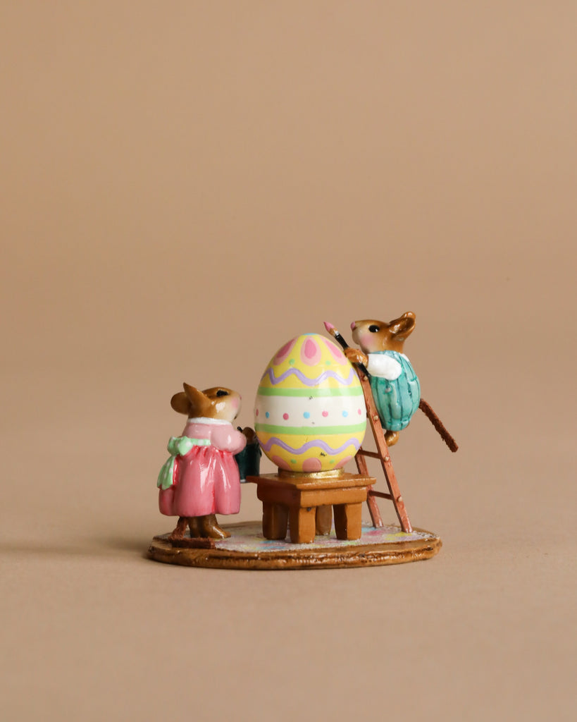 The Wee Forest Folk Tiny Collectible - Colossal Easter Coloring features two hand-sculpted mouse figurines, one in pink below and one in blue on a ladder, decorating a colorful Easter egg on a stand against a beige background.