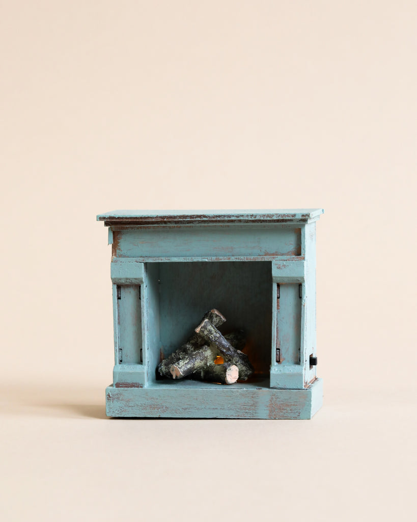 A charming Maileg Grandma And Grandpa Afternoon Tea wooden model fireplace in distressed light blue with faux logs inside, ideal for Maileg Mice, displayed against a simple beige background.