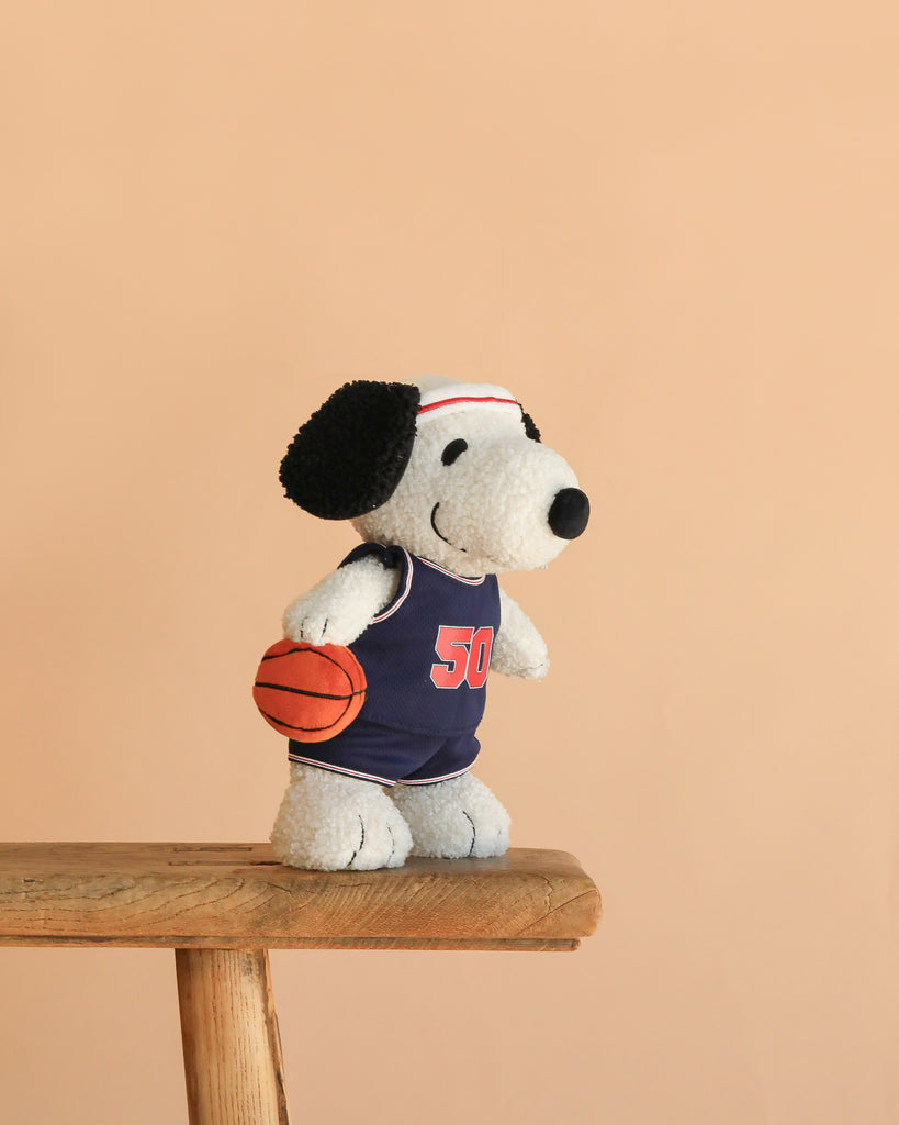 Peanuts Snoopy - Basketball plush, featuring Snoopy in a basketball jersey and headband, holding a basketball while standing on a wooden bench against a plain beige background.