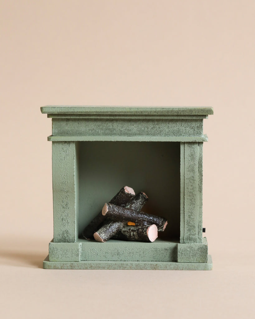 A small, green Maileg Miniature Fireplace with a vintage paint finish features four wooden logs stacked inside. The background is a plain beige color, emphasizing the minimalist and rustic design perfect for a Maileg House of Miniature.