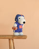The Peanuts Snoopy - American Football plush features Snoopy dressed as a football player in a blue #50 jersey and helmet, holding a football, standing on a wooden bench against a plain beige background.