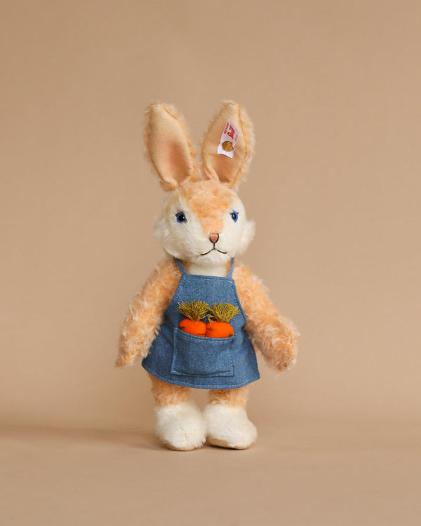 A plush toy Steiff, Carrie Springtime Easter Rabbit, Limited Edition Mohair, 8" bunny, standing upright, dressed in a denim dress holding a carrot, against a plain beige background. The bunny has long ears and a fluffy tail visible from the front.
