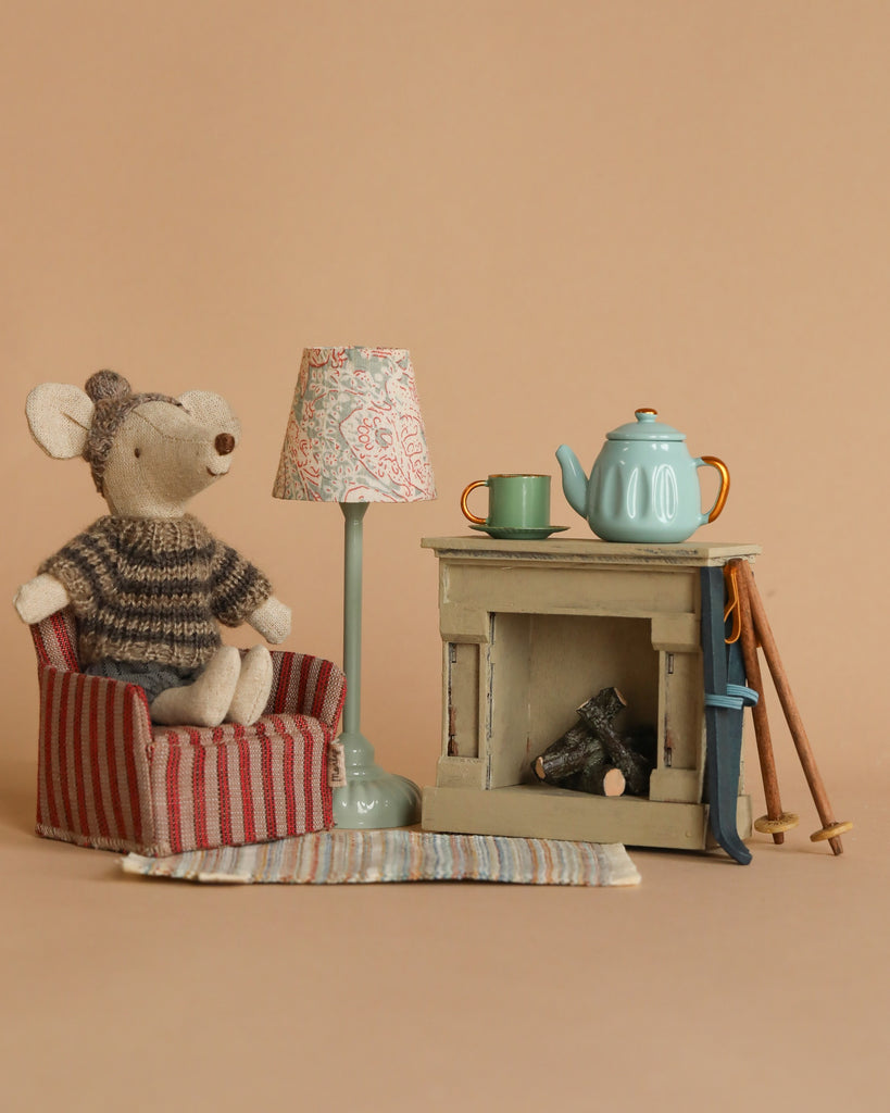 A cozy scene features the Maileg Winter Mouse with Ski Set, Big Brother - Grey, perched in a striped chair next to a patterned lamp. Nearby, a teal teapot rests with a cup on top. Ski poles suggest winter fun by the small fireplace filled with logs, and a striped rug adds warmth to the floor.