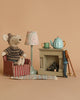 A cozy scene features a Maileg Chair, Mouse - Striped, occupied by a mouse doll beside a small fireplace resembling a gingerbread house. A colorful lamp, teapot, mug, and pair of walking sticks add to the charming setup alongside its Maileg friends, all set against a neutral background.