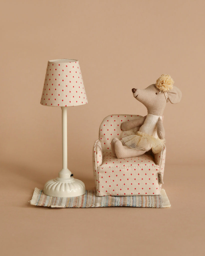 A small stuffed mouse in a tutu, one of the charming small Maileg friends, rests on a Maileg Chair, Mouse - Dot beside a matching lamp. The scene unfolds on a cream-colored background with a striped rug beneath the cozy setup, evoking the warmth of a gingerbread house.