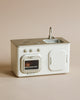 The Maileg Little Chef's Kitchen features a stovetop, oven, sink, and cabinet in minimalist white and silver, set against a beige background. Ideal for pairing with your Chef Mouse or Maileg Fridge accessories.