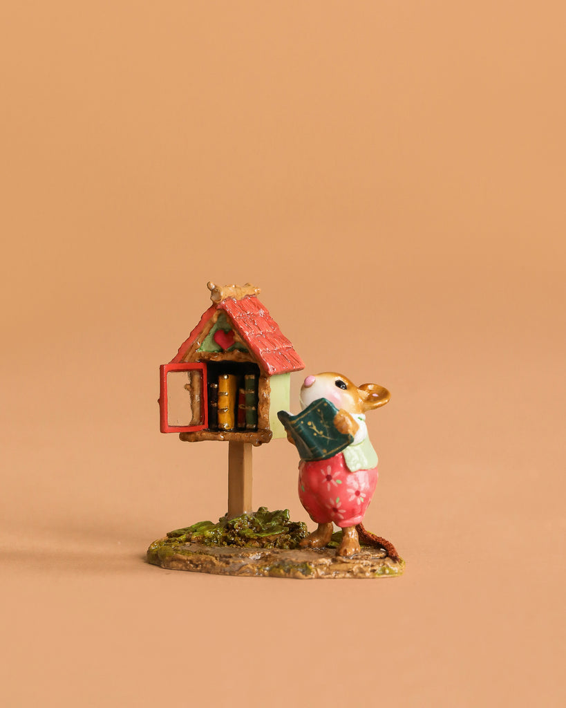 The Wee Forest Folk Tiny Collectible - Little Library in Pink features a mouse in a pink dress holding a blue book beside a red-roofed outdoor bookshelf, set against a beige background.