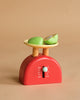 A red Kitchen Weighing Scales toy with a cream tray holds two green apple slice toys, displayed against a plain beige background.