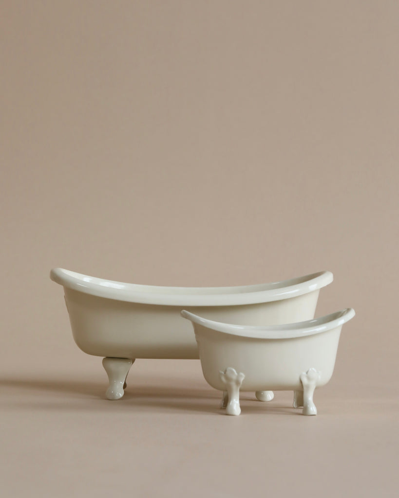 A minimalist beige background showcasing two small, white porcelain bathtubs. The larger vintage bathtub is to the left, while the smaller one is to the right. Both have a classic clawfoot design, and they're positioned side by side. Ideal for any Wellness Mouse in their own Maileg Bathtub (Miniature).