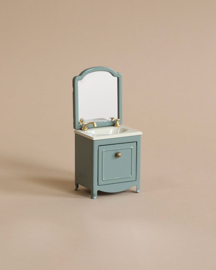 A Maileg Miniature Sink With Mirror with a removable round mirror and a single drawer, set against a plain beige background. The sink is gray with golden handles.