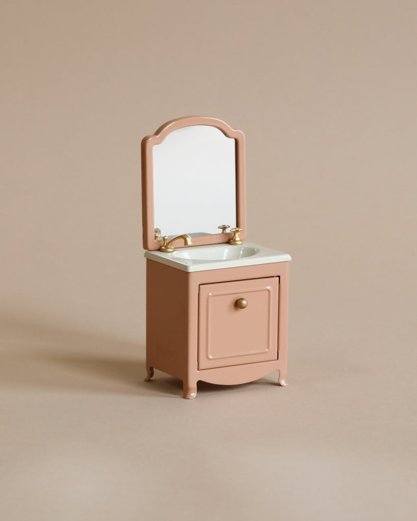 The Maileg Bonus Room Bathroom Set features a small, vintage-style pink vanity with a mirror, gold faucets, and a single cabinet door—perfect for enhancing any miniature dollhouse accessories collection. Set on a plain beige background.