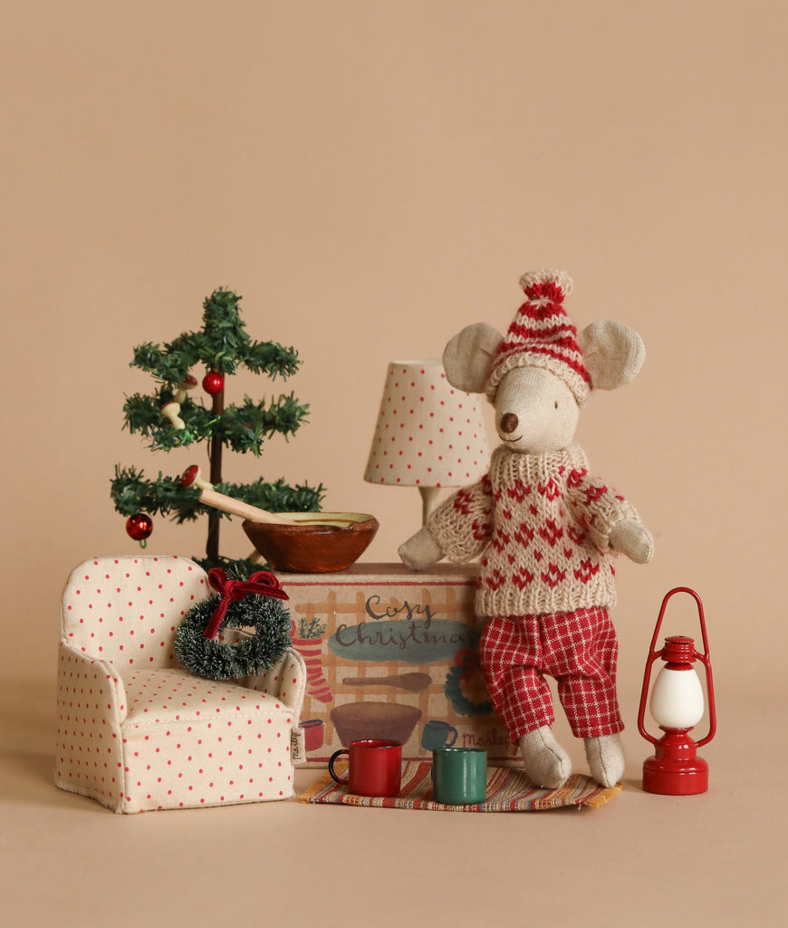 A cozy winter toy scene featuring the Maileg Winter Mouse with Ski Set, Mom - Red, dressed in a festive red and white knitted outfit. She stands next to a charming small polka-dotted armchair, a mini Christmas tree adorned with holiday decorations, as well as a red lantern and two mugs placed on a striped mat.