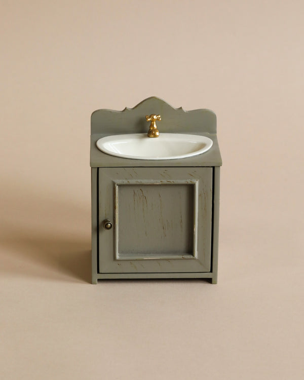 A Maileg miniature gray wooden bathroom vanity with a white basin and golden water tap, set against a plain beige background.