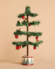 A small, minimalist Christmas tree from the Maileg Christmas Set features sparse green branches adorned with a few red and white ornaments, standing on a decorative base with geometric patterns reminiscent of a gingerbread house, set against a plain beige background.