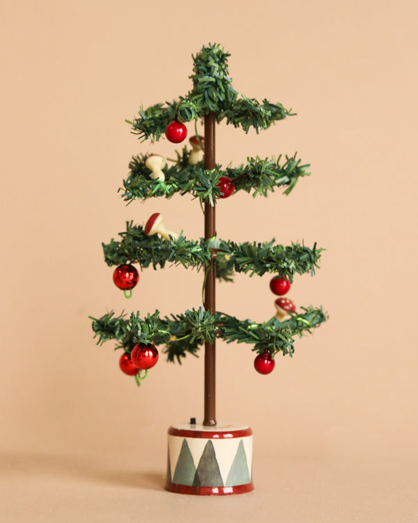 A small Maileg Christmas Tree from the Mouse collection features sparse branches decorated with red ornaments, small wooden figures, and twinkling LED bulbs. It stands on a circular base embellished with painted geometric patterns against a plain beige background.