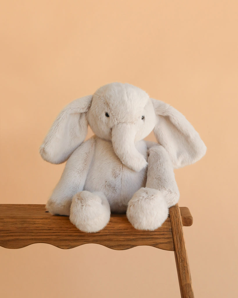 Snuggle Bunnies - Olive The Elephant, a soft plush with floppy ears, sits on a wooden surface against a peach backdrop—perfect as a cuddly toy for snuggles.