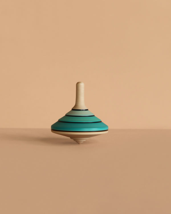 The Mader Tango Spinning Top - Blue, crafted from maple wood with blue, teal, and black stripes, stands upright on a beige surface against a matching beige background.