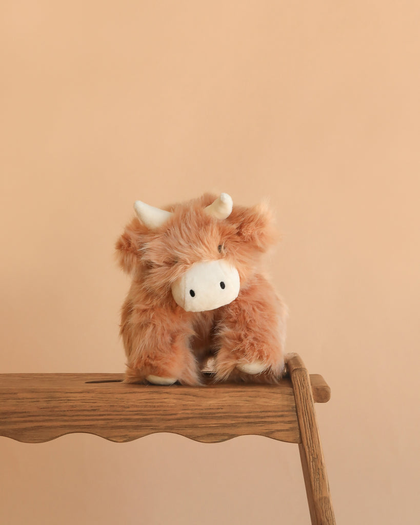 Snuggle Bunnies - Hilda The Highland Cow - Caramel is a soft, light brown plush with small horns and a white snout, shown on a wooden stool against a beige background—a charming cuddle buddy or baby shower gift.