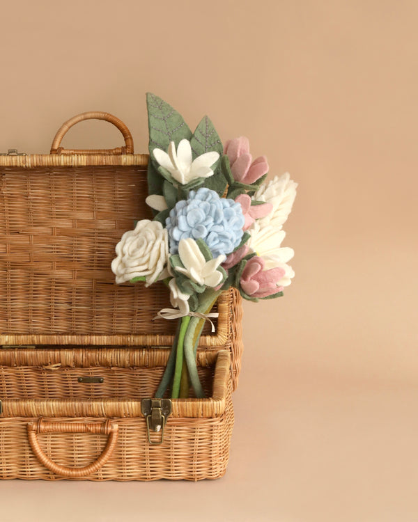 A vintage wicker picnic basket with a handle and the Felt Flower Bouquet - Muted Blooms peeking out against a muted beige background.