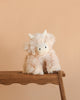 Snuggle Bunnies - Hilda The Highland Cow, a fluffy light beige plush with white horns, sits on a wooden bench against a peach background—the perfect cuddle companion.