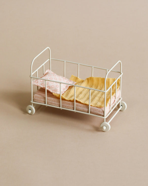 A Maileg Micro Cot bed with wheels, adorned with pink and beige patterned bedding, set against a neutral beige background.