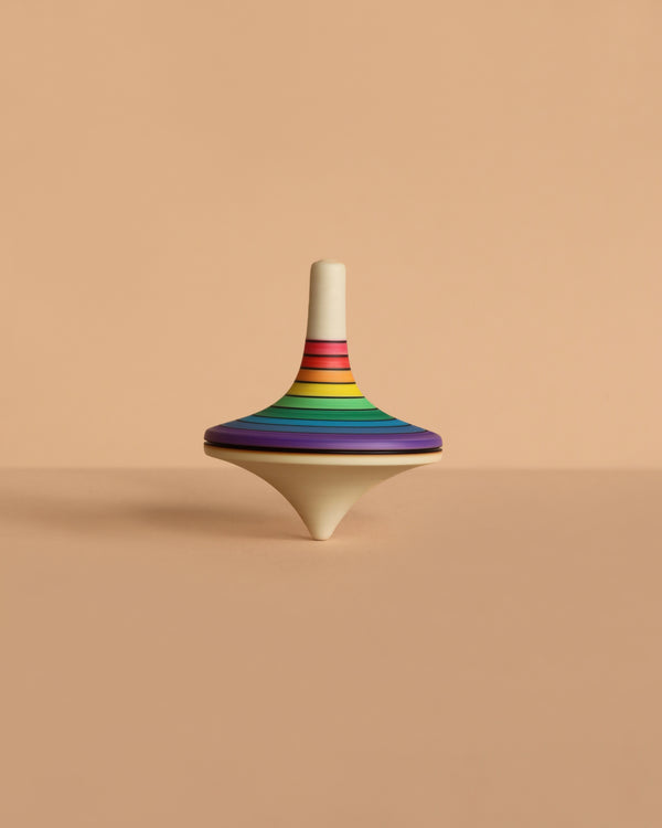The Mader Large Spinning Top - Rainbow, featuring vibrant stripes, stands upright on a beige surface against a matching beige background.