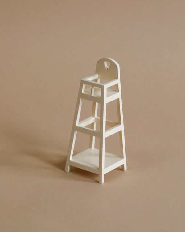 A small, white wooden Maileg My High Chair sits on a beige background. The nursery furniture piece has a simple design with a cutout detail on the backrest and no additional adornments.