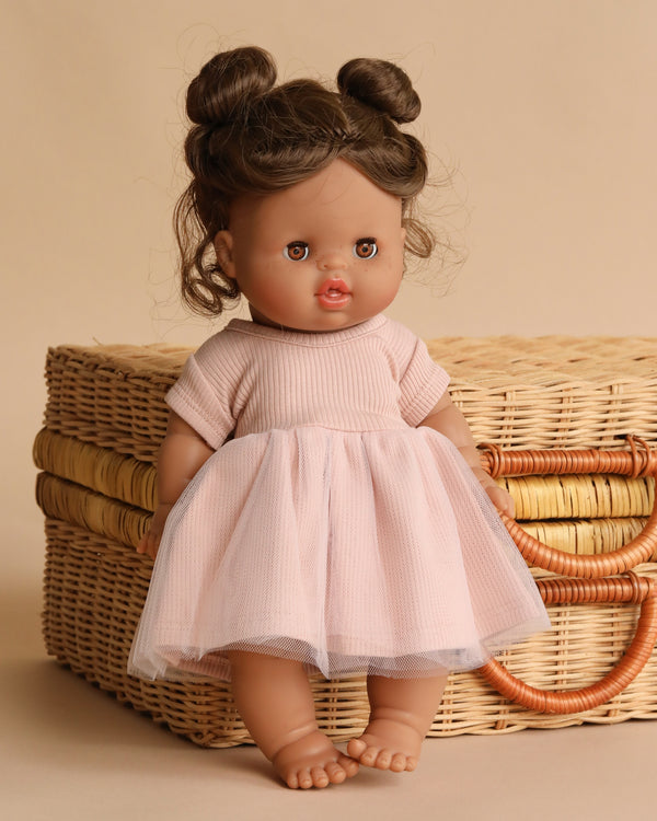 The Minikane Doll With Sleeping Eyes (13") - Madeleine with Clothing stands barefoot in front of a woven wicker basket, wearing a light pink dress, her brown hair styled in two buns, set against a beige background.