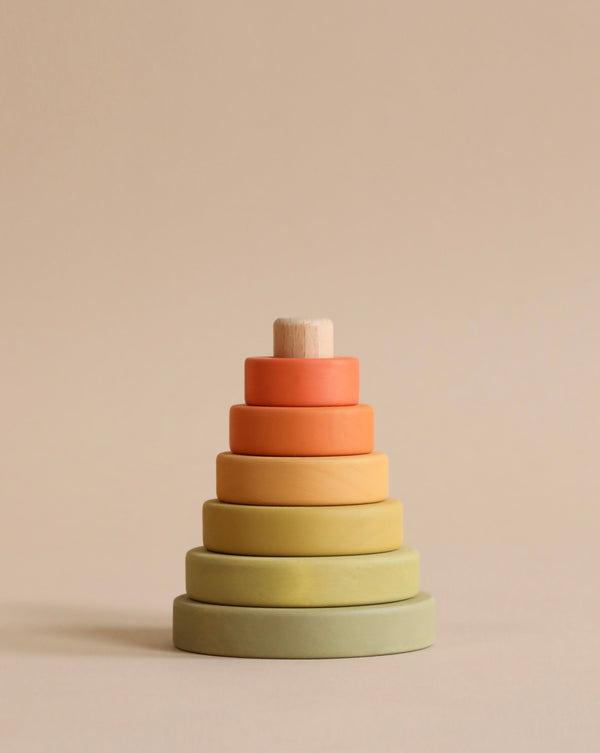 A stack of Mini Wooden Pyramid Stacker - Flower Meadow coated in non-toxic paint, in a gradient of warm tones, from dark green at the base to soft peach at the top, set against a neutral beige background.