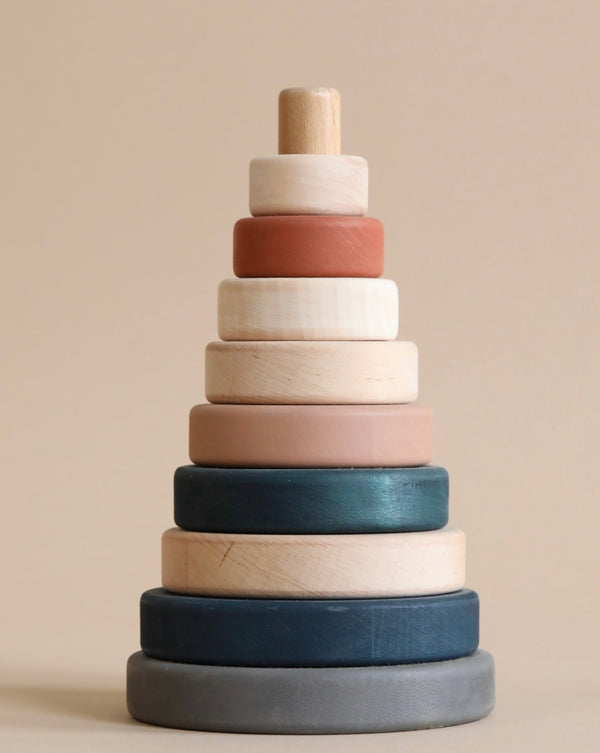 A Wooden Pyramid Stacker - Terracotta featuring rings of varying sizes, painted with non-toxic paint in different muted colors, arranged in order from largest at the bottom to smallest at the top, against a beige background.