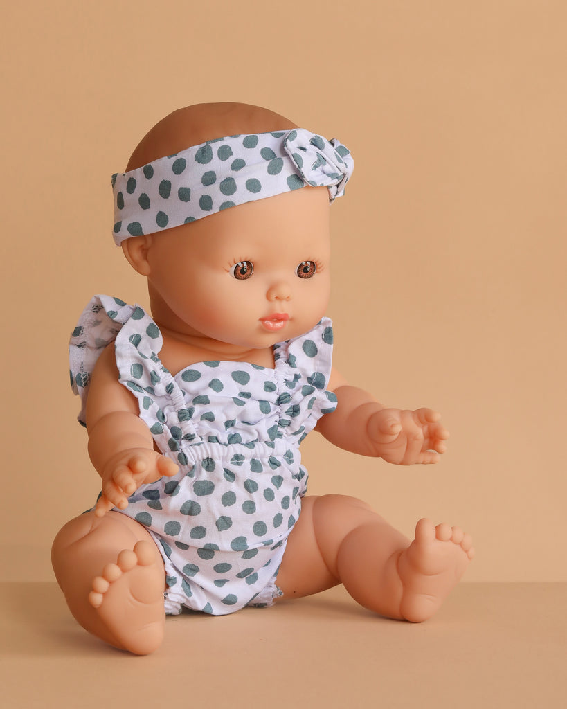 The Minikane Doll (13") - Faustine with Clothing wears a white outfit with green polka dots and a matching headband. Made in Spain from phthalate-free vinyl, it's perfect for little hands and sits on a light brown surface against a beige background.