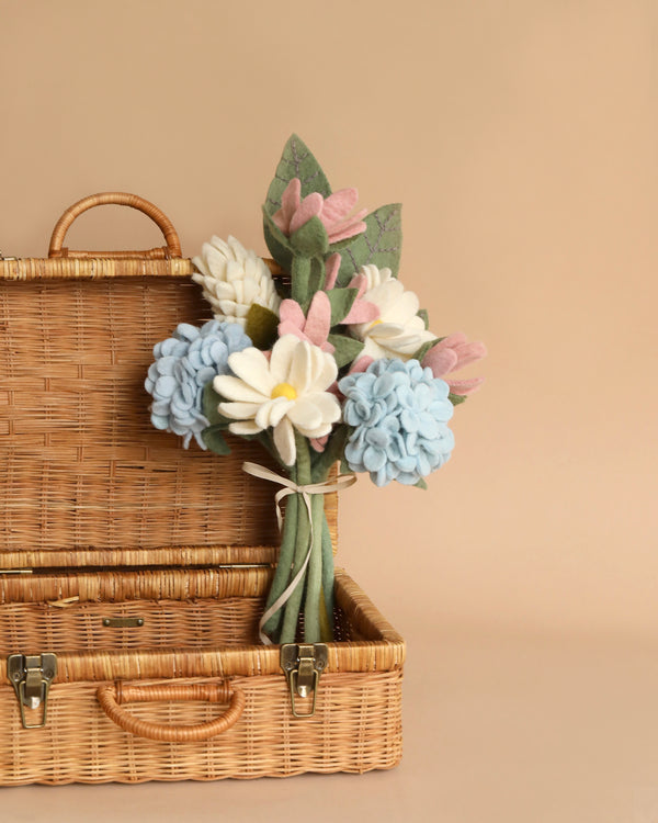 A handmade in Nepal picnic basket with a handle, partially opened, revealing a Felt Flower Bouquet - Blooming Meadow made up of white, blue, and pink flowers against a soft beige background.