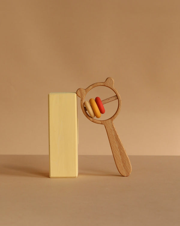 A wooden block stands upright next to the Sabo Concept Bear and Rings Rattle—featuring a bear-shaped handle and three rings in yellow, orange, and pink—against a beige background.
