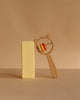 A wooden block stands upright next to the Sabo Concept Bear and Rings Rattle—featuring a bear-shaped handle and three rings in yellow, orange, and pink—against a beige background.