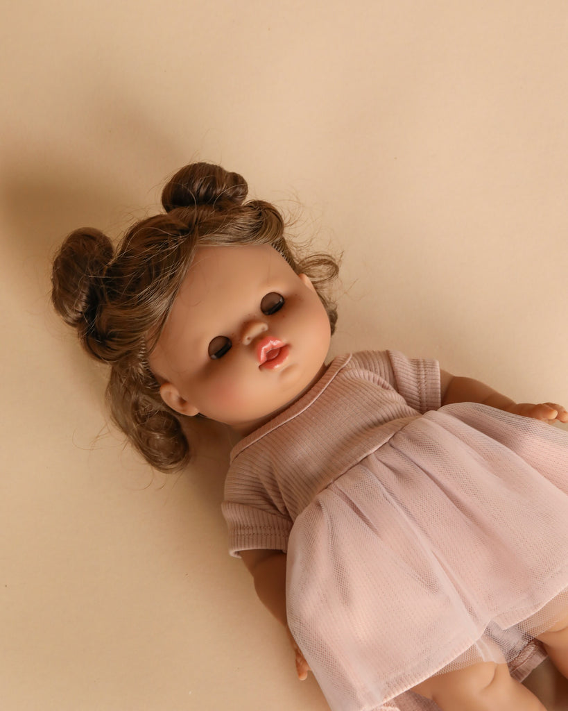 The Minikane Doll With Sleeping Eyes (13") - Madeleine with Clothing, featuring brown hair in two buns and a pink dress, rests on a beige surface.