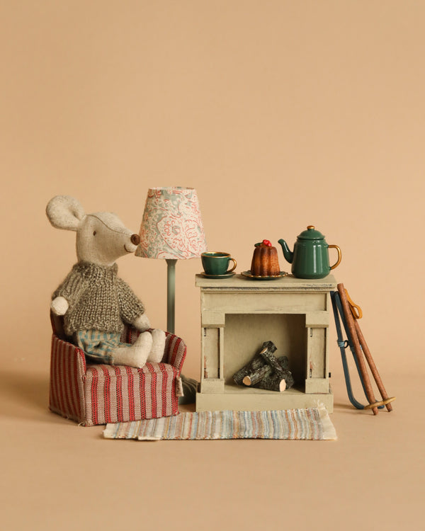 The Maileg Ski Lounge Set features a small stuffed children's toy called Big Brother Ski Mouse, who is dressed in a knit sweater and seated on a striped armchair next to a miniature fireplace. Adjacent to them are a green teapot, cup, and saucer on the mantel, along with a standing lamp, skis, and a striped rug. The entire cozy scene is set against a soft beige background.