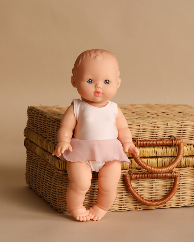 The Minikane Vintage Baby Doll (13") - Rosalie with Clothing, made in Spain, stands in front of two stacked wicker suitcases against a beige background.