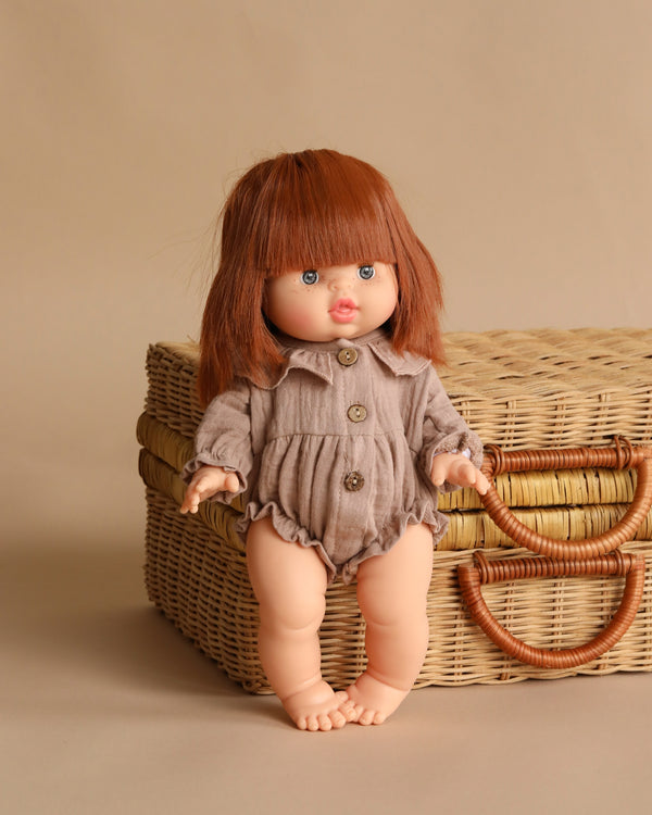 The Minikane Baby Doll (13") - Capucine with Clothing features straight auburn hair and bangs, a taupe buttoned romper, and stands barefoot in front of a wicker suitcase with handles against a beige background.
