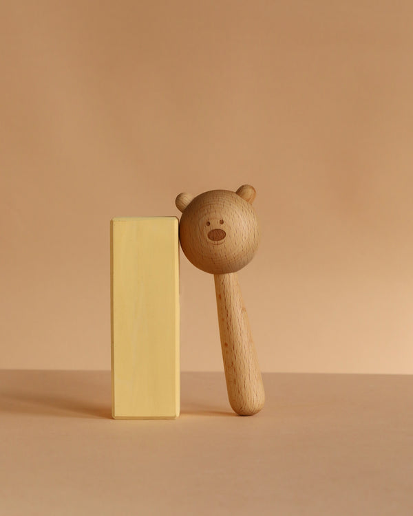 The Sabo Concept Bear Rattle - Natural, beautifully crafted by a renowned Ukrainian brand, features a round head and simple face, leaning on a rectangular yellow block against a beige background.