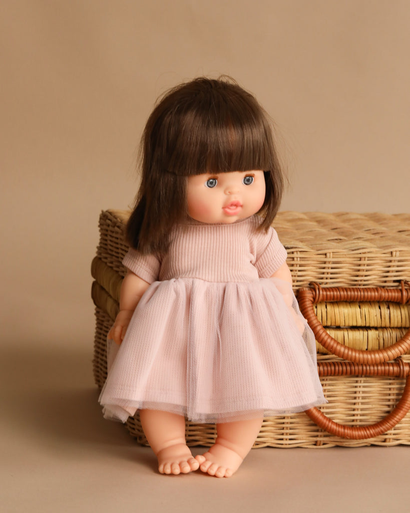The Minikane Baby Doll (13") - Chloe with Clothing stands barefoot in front of a woven wicker basket, wearing a light pink dress. She has straight brown hair and blue eyes, set against a beige background.