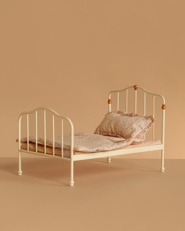 The Maileg Vintage Bed, Mouse Size - Off White features a simple vintage design with curved headboard and footboard, a patterned pillow and blanket, and is perfect as charming dollhouse furniture. Set against a plain beige background.