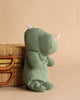 The Maileg Rhino, Medium - Sage, a soft green stuffed toy from the Safari Friends collection, is placed beside a wicker basket with leather handles against a neutral beige background.