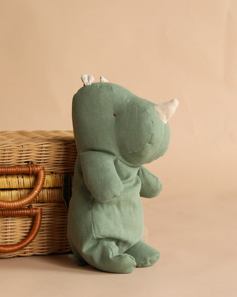 The Maileg Rhino, Medium - Sage, a soft green stuffed toy from the Safari Friends collection, is placed beside a wicker basket with leather handles against a neutral beige background.