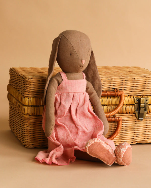 The Maileg Bunny, Size 4 - Brown, handmade from natural fabrics and linen, wears a pink dress and shoes while sitting in front of a closed wicker suitcase against a beige background.