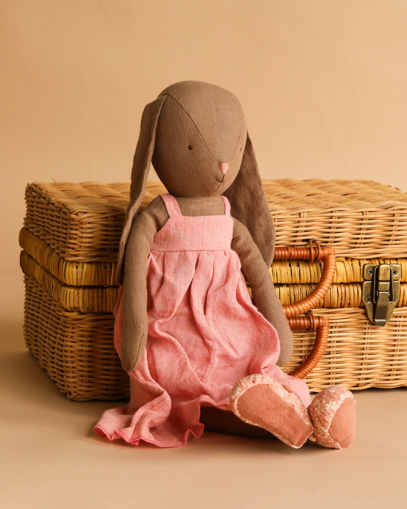 The Maileg Bunny, Size 4 - Brown, handmade from natural fabrics and linen, wears a pink dress and shoes while sitting in front of a closed wicker suitcase against a beige background.