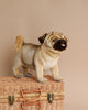 A realistic Pug Dog Stuffed Animal, part of the HANSA animals collection, standing on a woven wicker suitcase against a soft beige background. The toy features detailed facial wrinkles and a curly tail.